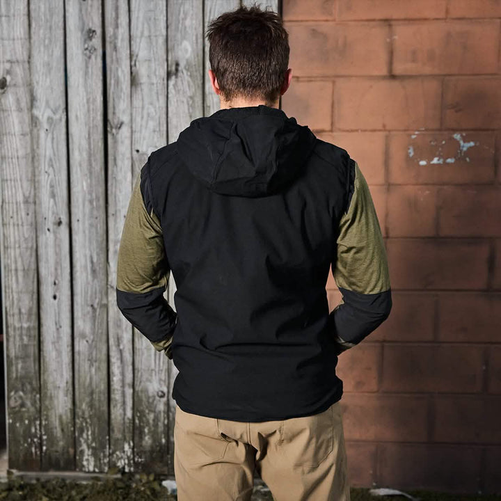 A man wearing the Vest of Power - ToughDry® + Fleece by GORUCK and khaki pants stands with his back to the camera, hands in pockets, capturing the environmentally conscious spirit of outdoor adventure. Positioned against a wooden fence and brick wall, he effortlessly merges casual style with mindful living.