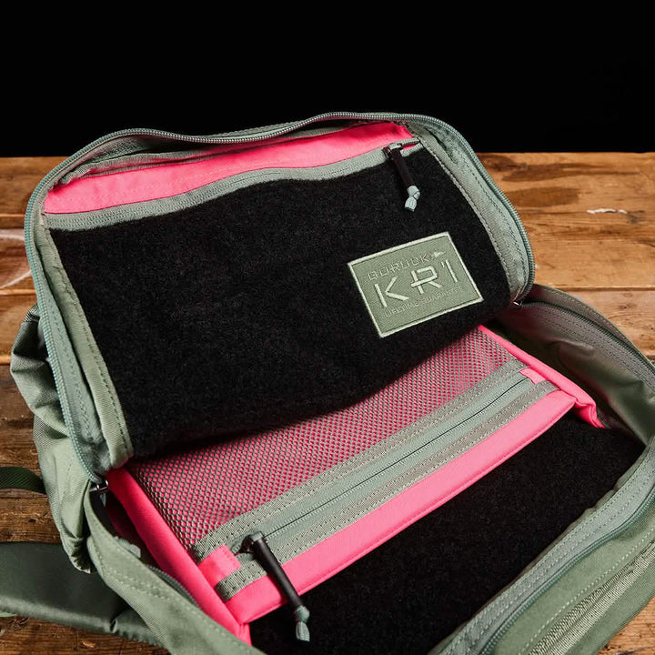 An open KR1 kids rucksack from GORUCK rests on a wooden table, showcasing its pink interior compartments with a mesh pocket and a patch labeled KPR affixed to the black surface. This green backpack, ideal for carrying school supplies, also boasts padded straps for enhanced comfort.