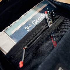 Close-up of a black zippered mesh pocket inside a GORUCK bag holding a 3rd Grade workbook and pens