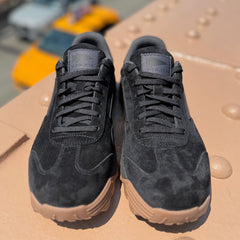 A pair of Mackall 45 Black + Gum sneakers with black suede uppers and tan soles are placed on a textured beige surface outdoors.