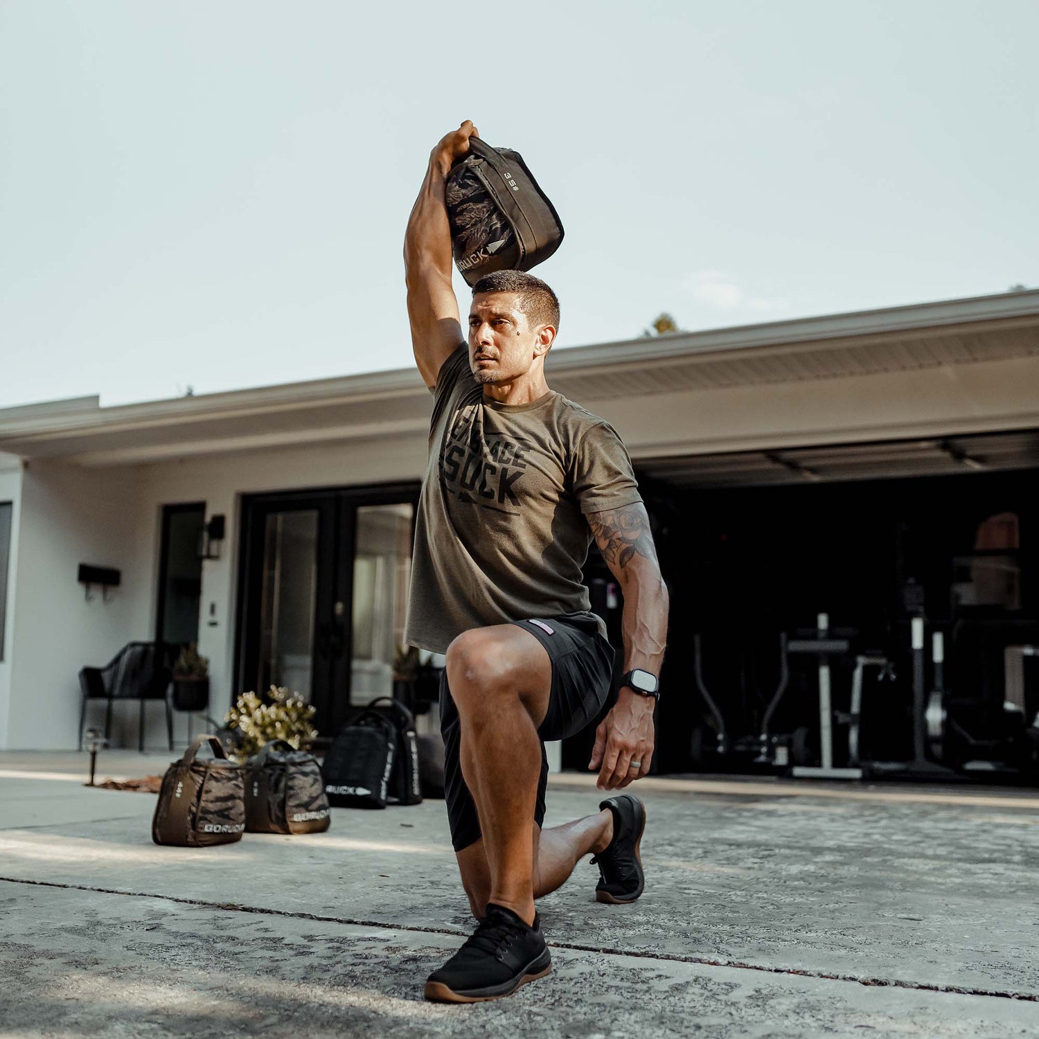 A man lunges outdoors, holding a Sand Kettlebell overhead with gym gear nearby, demonstrating the effectiveness of at-home workouts using portable Sand Kettlebells.