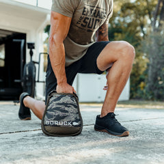 A man in athletic wear kneels outdoors, holding a Sand Kettlebell with a camo pattern—ideal for at-home training or workouts on the go.