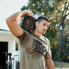 Wearing a shirt that says "Embrace the Suck," a man in workout clothes lifts Sand Kettlebells over his shoulder outdoors. This portable kettlebell alternative is perfect for at-home workouts and building strength anywhere.