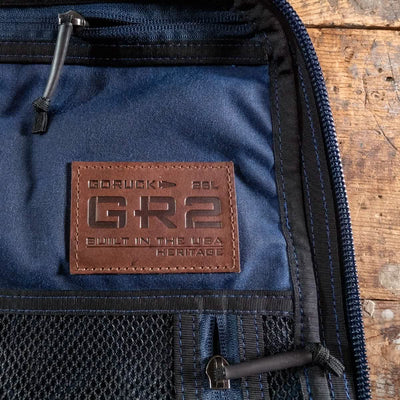 Close-up of a blue waxed canvas backpack featuring the GORUCK GR2 Heritage with an alligator leather front panel and a brown leather patch reading “Built in the USA.” The bag showcases black zippers and a mesh pocket against a wooden surface background.