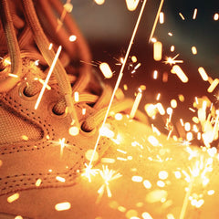Close-up of tan GORUCK MACV-2 Safety Boot - Mid Top with large sparks flying around them, suggesting metal welding or grinding. The bright orange and yellow sparks contrast with the textured surface of the boots, creating a dynamic and energetic scene.