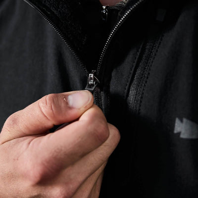 A person zipping up a black GORUCK Vest of Power made with ToughDry® and fleece, with focus on their hand gripping the zipper. This environmentally conscious design stands out against the dark background, emphasizing the action of zipping.