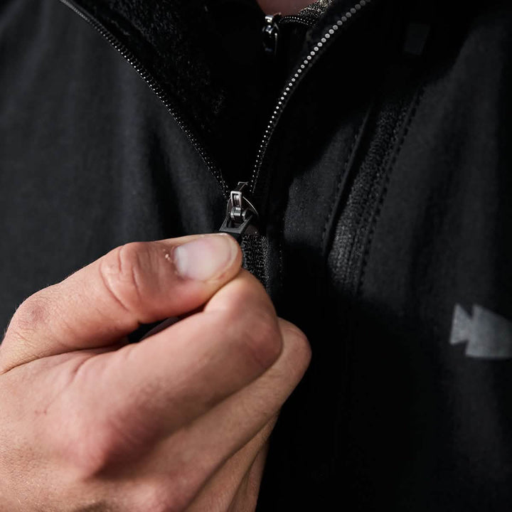 A person zipping up a black GORUCK Vest of Power made with ToughDry® and fleece, with focus on their hand gripping the zipper. This environmentally conscious design stands out against the dark background, emphasizing the action of zipping.
