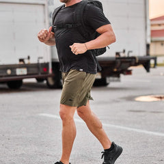 Man running outdoors wearing black shirt, green GORUCK shorts, black backpack, and black sneakers in urban setting