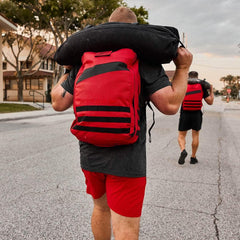Two men carrying black sandbags with GORUCK red rucking backpacks walking on a street at dusk