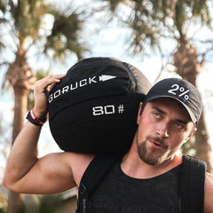 A man outdoors, ideal for a workout focused on core stability, is carrying a black GORUCK Sand Medicine Ball marked with 80# on his shoulder. He sports a black cap featuring the number 2% and wears a black tank top. In the blurred background, tall palm trees can be seen.