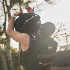 A person wearing a black cap and backpack is outdoors, lifting a heavy 80LB GORUCK Sand Medicine Ball, showcasing impressive core stability. The sun is shining, and trees surround this home gym-friendly workout setting.