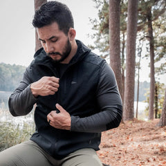 A man sits outdoors by a tranquil lake, embraced by nature's greenery. Dressed in the GORUCK Vest of Power made from ToughDry® + Fleece over a long-sleeve gray shirt and green pants, he thoughtfully adjusts the zipper. The serene, forested backdrop mirrors his eco-friendly mindset.