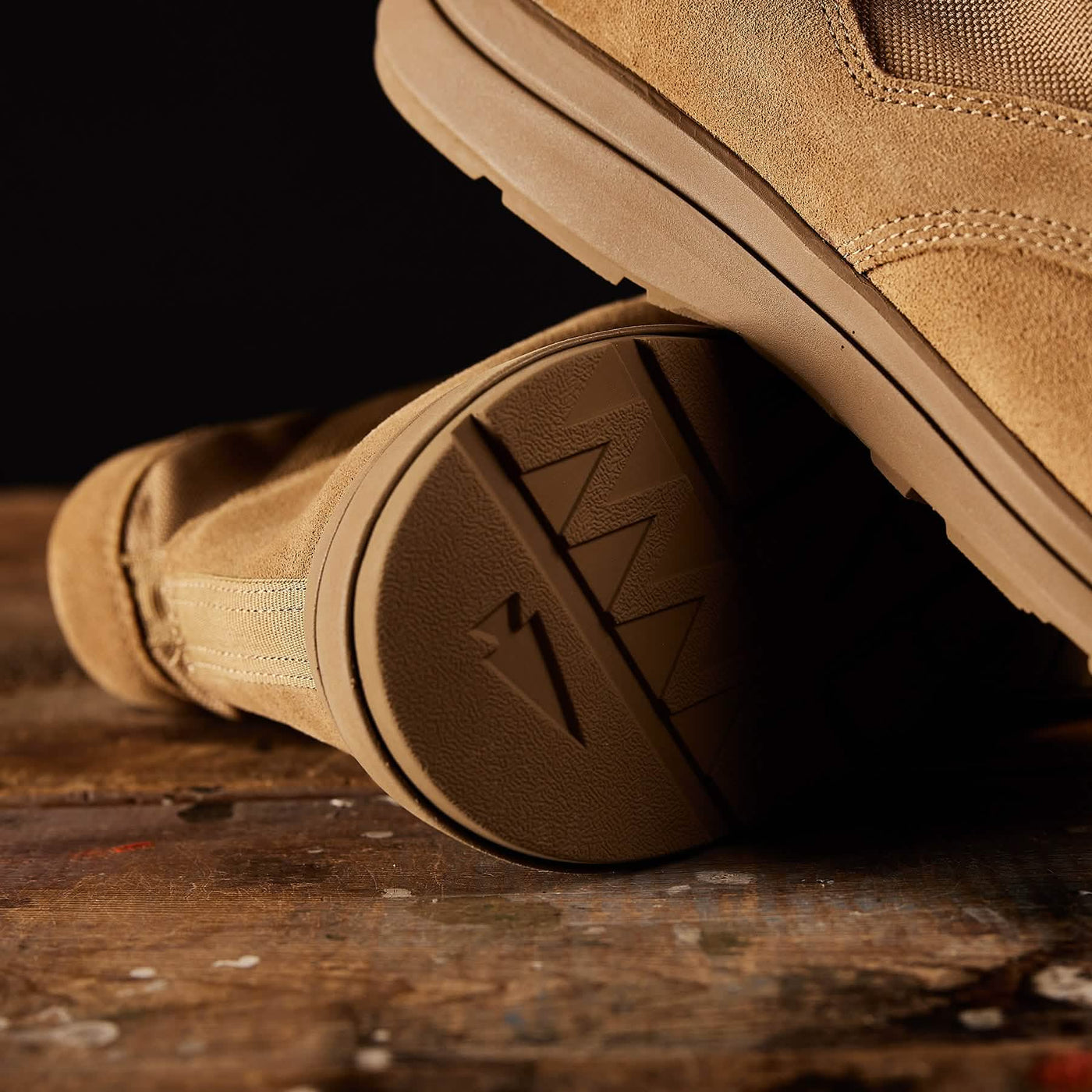 Close-up of a lightweight tan suede boot resting on a wooden surface. The focus is on the textured rubber sole, showcasing a distinctive zigzag pattern and a small embossed lightning bolt. Reminiscent of the MACV-1 Hi-Speed - Mid Top by GORUCK, the dark background highlights the details of these rugged rucking boots.