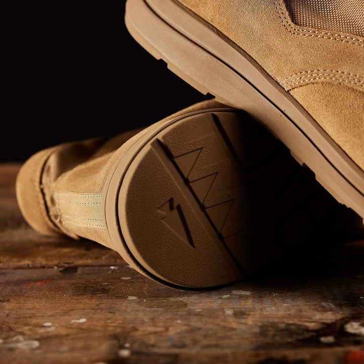 Close-up of a lightweight tan suede boot resting on a wooden surface. The focus is on the textured rubber sole, showcasing a distinctive zigzag pattern and a small embossed lightning bolt. Reminiscent of the MACV-1 Hi-Speed - Mid Top by GORUCK, the dark background highlights the details of these rugged rucking boots.