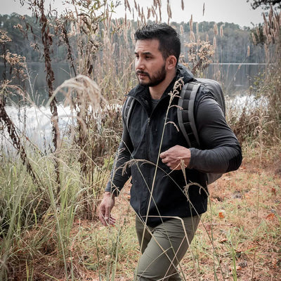 A man with a GORUCK Vest of Power - ToughDry® + Fleece hikes through tall grass beside a lake. He sports olive green pants as trees in the background frame the shimmering water, creating an environmentally conscious ambiance that is both calm and natural.