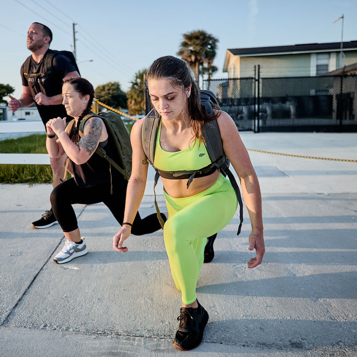 Three people exercise outdoors with heavy bags. A woman lunges in front, wearing bright yellow GORUCK Women’s Training Leggings made of squat-proof ToughFlex fabric, providing comfort and durability for intense workouts.