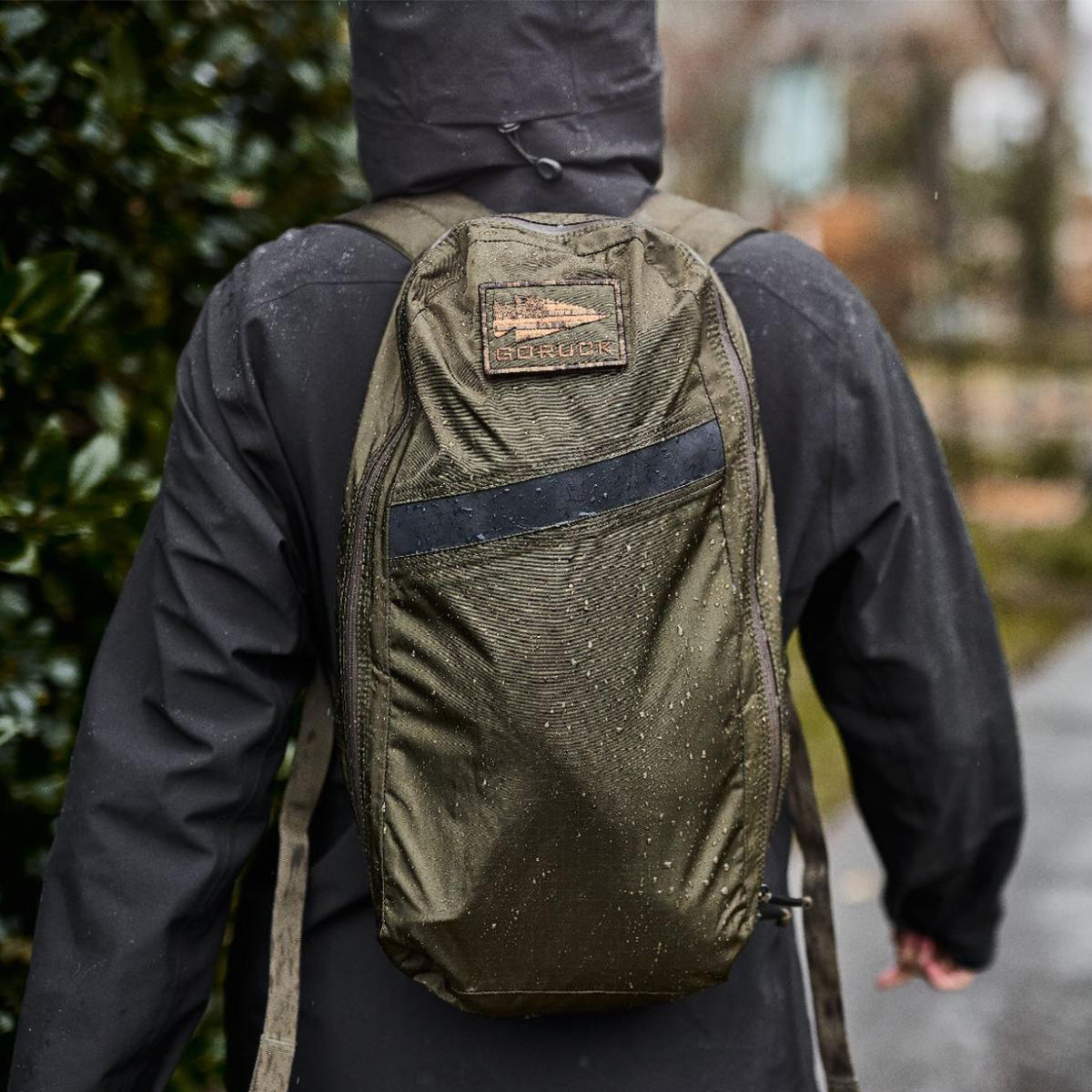 Person in a dark rain jacket wearing an olive green GORUCK backpack outdoors on a wet day.