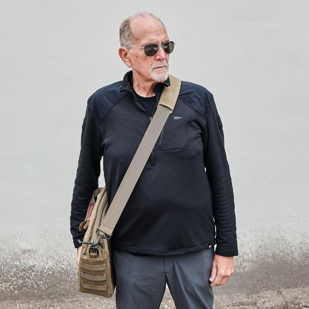 Older man wearing sunglasses and a black top, carrying a tan shoulder bag, standing against a plain wall.