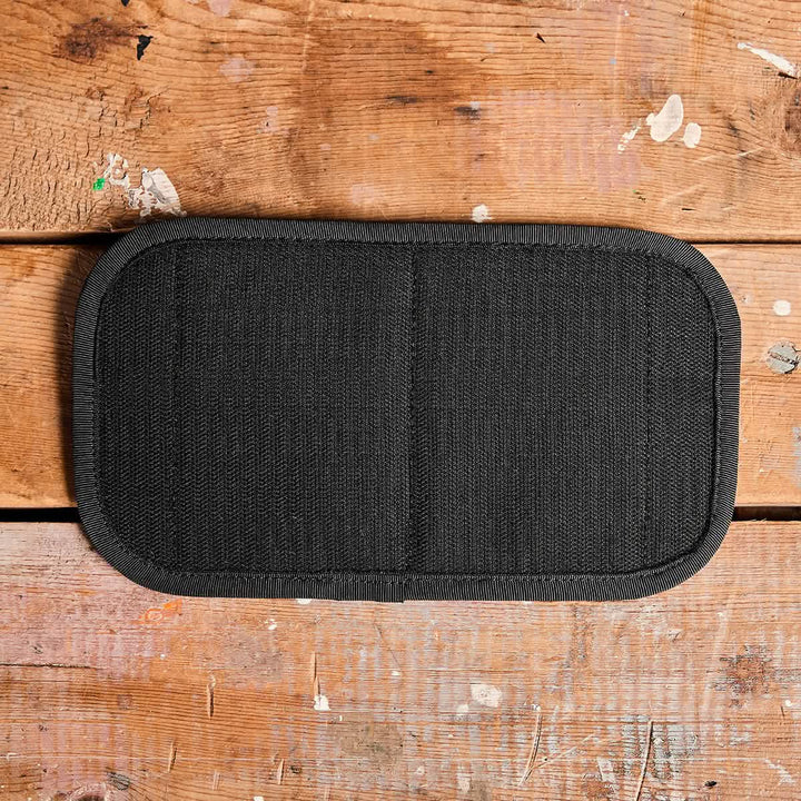 An Admin Panel by GORUCK, ideal for EDC enthusiasts or rucking adventures, lies on a worn wooden surface with scattered paint marks and scratches. The panel is black with a rectangular shape, rounded edges, and stitching down the center.