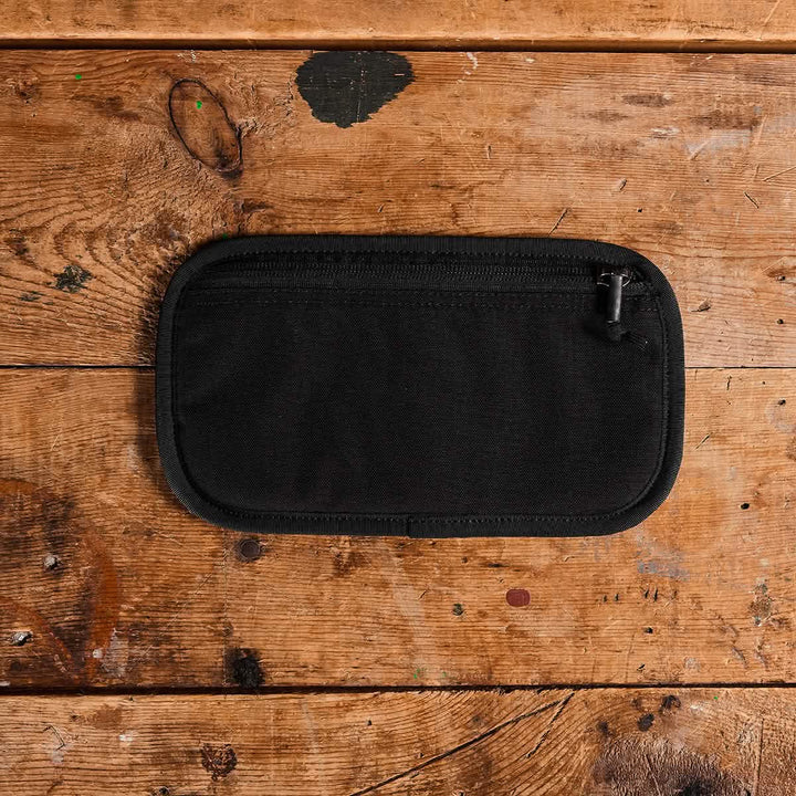 A compact, rectangular black Admin Panel by GORUCK sits flat on a rustic wooden surface, ideal for EDC. The wood features various stains and knots, contributing to the rugged texture of the background.