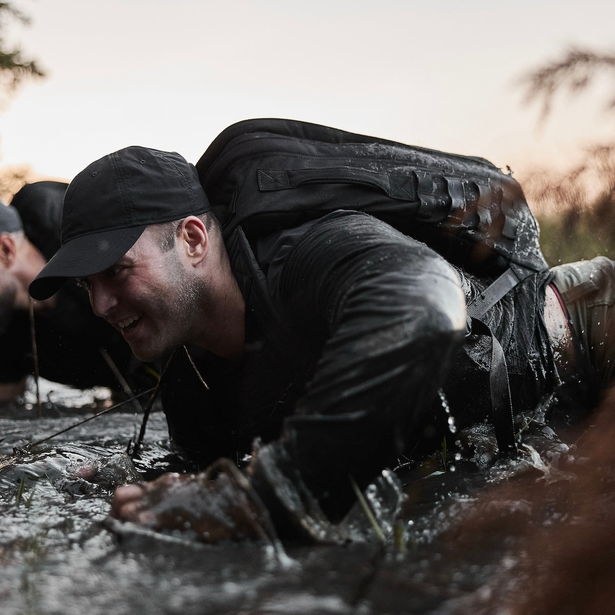 Man in dark clothing and cap crawls through muddy water, smiling, during an outdoor obstacle course.
