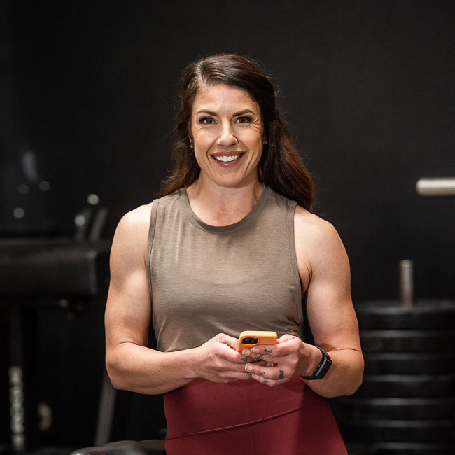Smiling woman in athletic wear holds a phone in a gym with weights and equipment in the background.