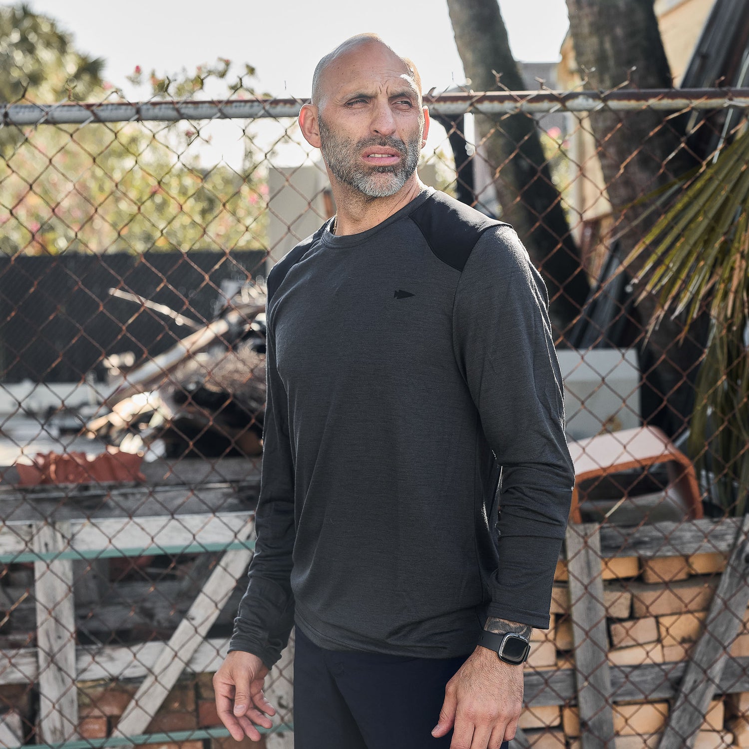 A bald man with a beard stands outside by a chain-link fence, wearing the Men’s Commando Long Sleeve - Merino Wool top.
