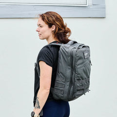 A person with reddish-brown hair stands sideways, carrying a large gray GR2 - Cordura ruck by GORUCK. They wear a black T-shirt and have a tattoo on their left forearm, set against a plain light-colored wall, exuding an air of understated strength reminiscent of Special Forces precision.
