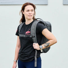 A person with shoulder-length hair stands against a light wall, wearing a black t-shirt featuring a red graphic and carrying the GORUCK GR2, known for its carry-on compliant design and Special Forces quality. They have a tattoo on their left forearm and are looking to their right.