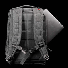 A black GORUCK GR2 backpack with padded shoulder straps and a quilted back sits against a black background. A laptop is partially visible, either being inserted or removed from the side pocket. The scene is well-lit, emphasizing the backpack's texture, details, and its Special Forces quality design.