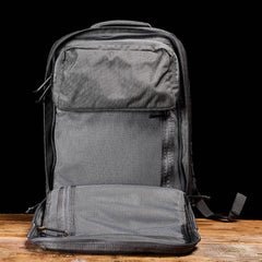 The GORUCK GR2 backpack, featuring multiple compartments such as a front pocket and a zippered section, is showcased in gray against a black background on a wooden surface. Designed to meet Special Forces quality standards, it offers excellent durability and carry-on compliant convenience for those who seek reliability.