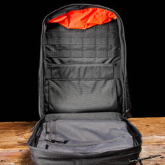 The GR2 - Cordura by GORUCK, an open black backpack, reminiscent of those used by Special Forces, sits on a wooden table, showcasing its spacious interior. With an orange lining, multiple compartments, and a mesh pocket for organizing essentials, it highlights its impressive storage capacity against a plain black background.