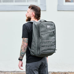 A man with a tattooed arm stands outdoors in front of a light-colored building, wearing a large gray GR2 - Ballistic Nylon Cordura backpack.