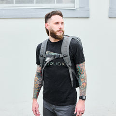 A bearded man with tattooed arms wears a black GORUCK shirt and stands against a light wall, carrying the GR2 - Ballistic Nylon Cordura rucksack, an ideal carry-on travel bag.