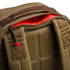 Close-up of the Double Compartment Bullet Ruck - Heritage Waxed Canvas - 18L, featuring padded straps and a khaki waxed canvas exterior with a zippered pocket lined in bright red fabric.