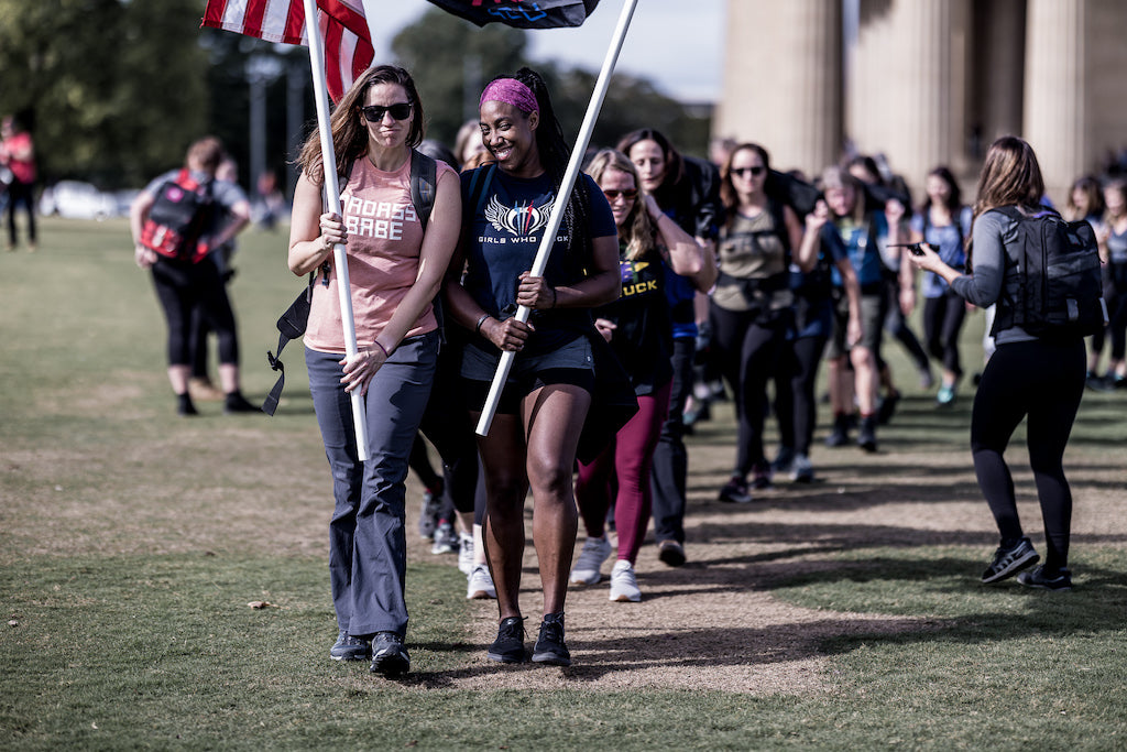 Two women holding flags lead a group of people walking outdoors on a grassy path near columns.