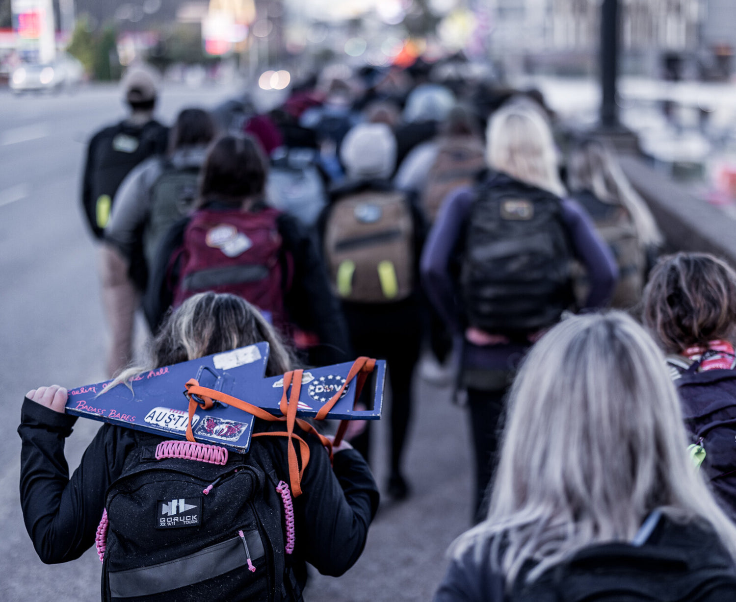 A group of people with backpacks walk along a street; one person carries a decorated blue board.