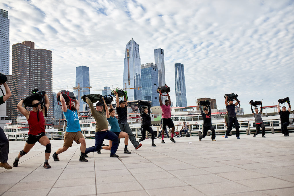 Group of people exercising outdoors with sandbags, skyscrapers in the background, cloudy sky above.