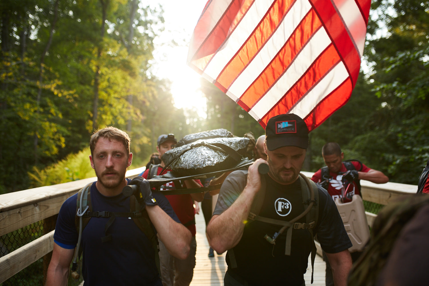 Group of men carrying a stretcher and an American flag while walking on a bridge in a forested area.