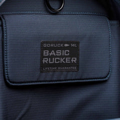 Close-up of the front pocket on a blue Basic Rucker backpack, featuring a stitched label and designed for comfortable rucking with secure storage for your ruck plate.