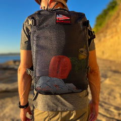 A person carries the Mesh Ruck, a black mesh backpack with visible gear inside, while walking outdoors on a sunny day.