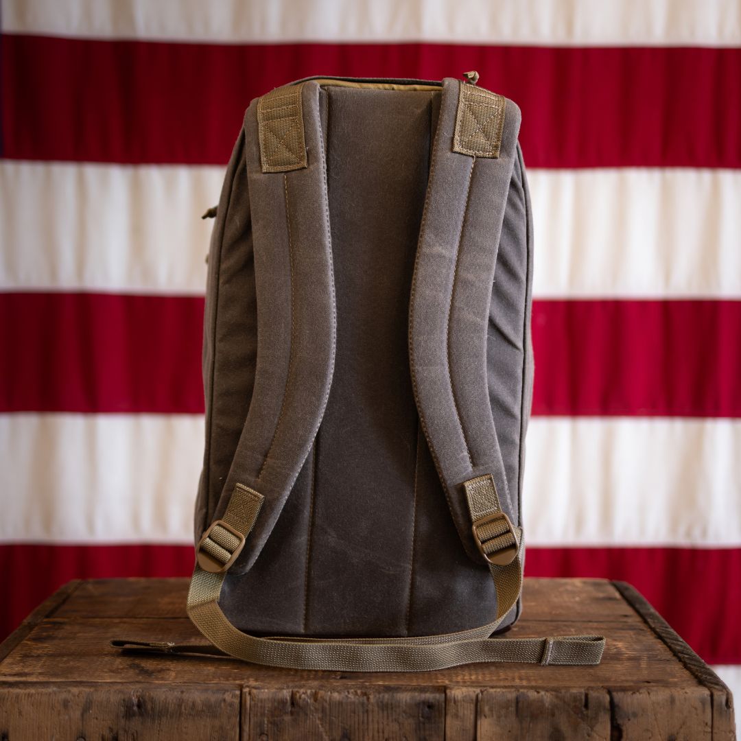 Sashiko Waxed Bullet - Built by Scars with padded straps rests on a wooden table, flag in the background.