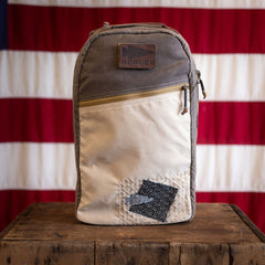 A tan and gray Sashiko Waxed Bullet - Built by Scars backpack sits on a wooden box, US flag in background.