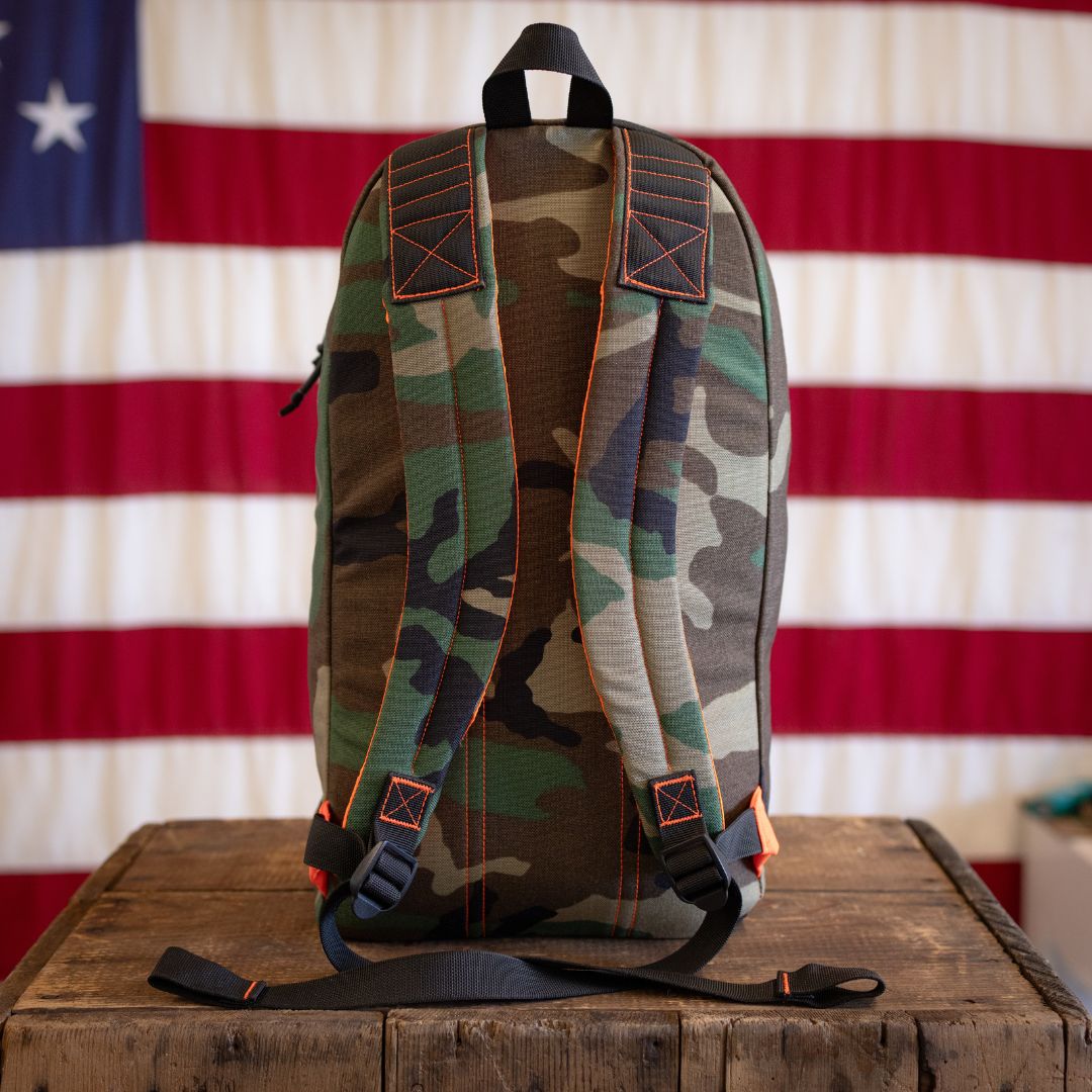 Woodland Camo 15L Bullet backpack with orange stitching and black straps sitting on a wooden crate in front of an American flag.