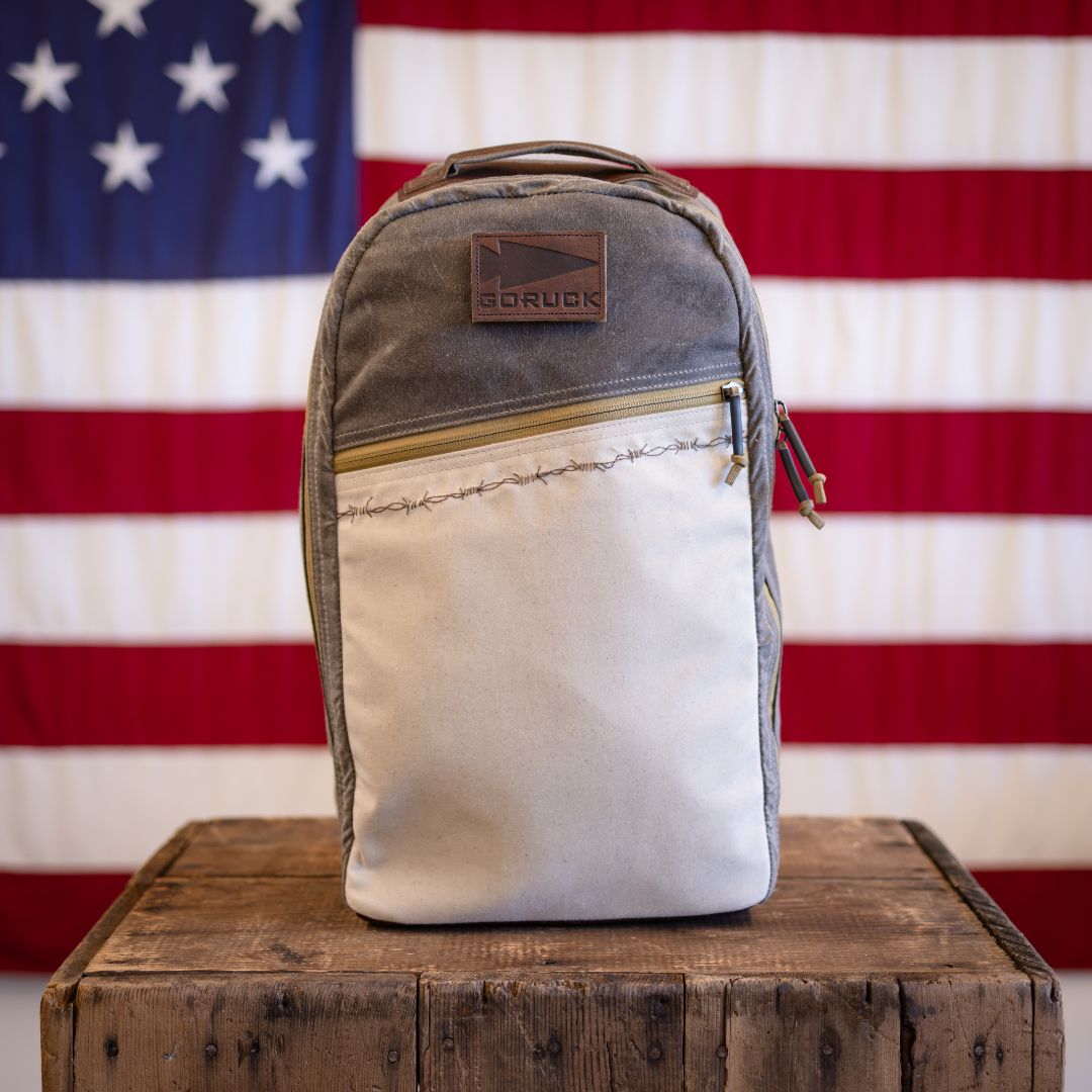 A two-tone, limited edition GORUCK backpack with a leather logo patch sits on a wooden crate in front of an American flag.