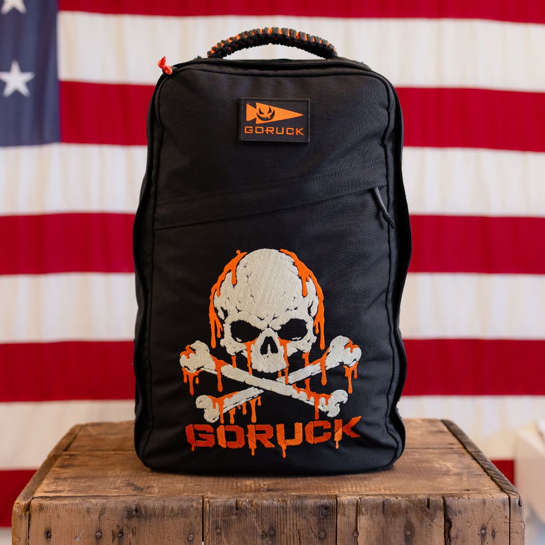 Black limited edition GR1 rucksack featuring an orange dripping skull and crossbones graphic, sitting on a wooden crate in front of an American flag.