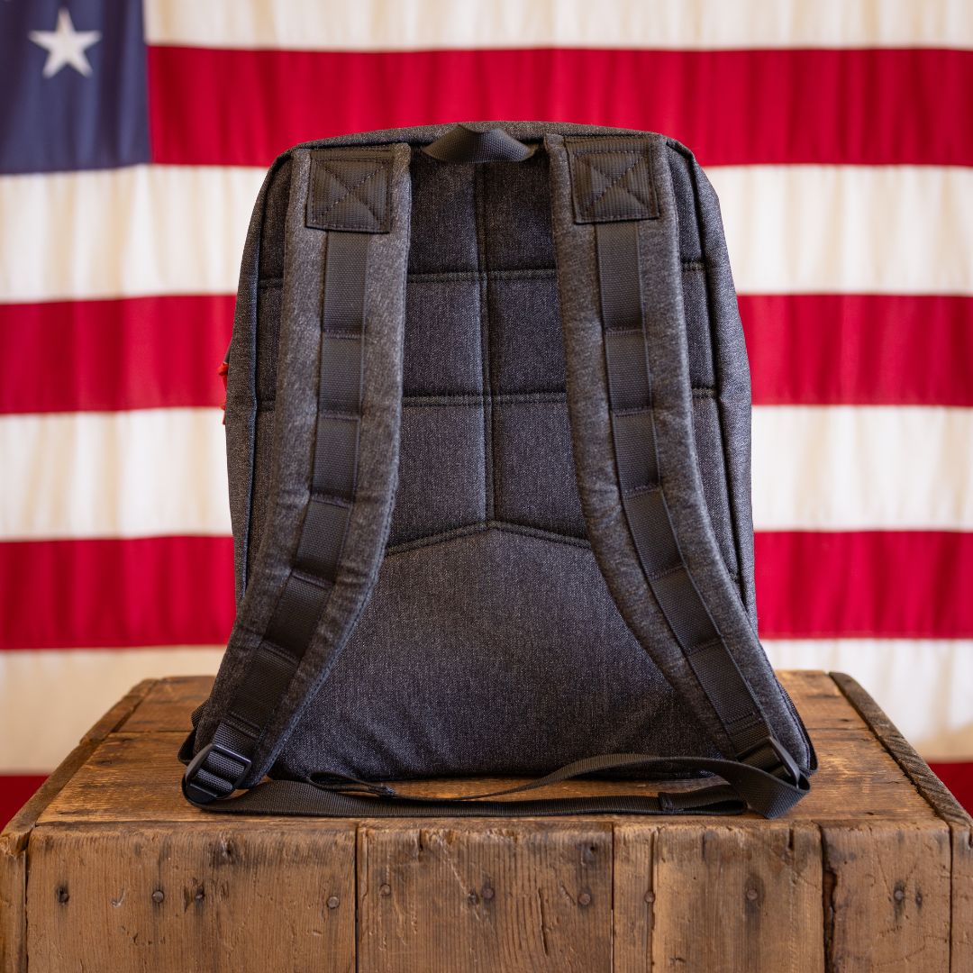 A handmade charcoal grey backpack sits on a wooden crate in front of an American flag.