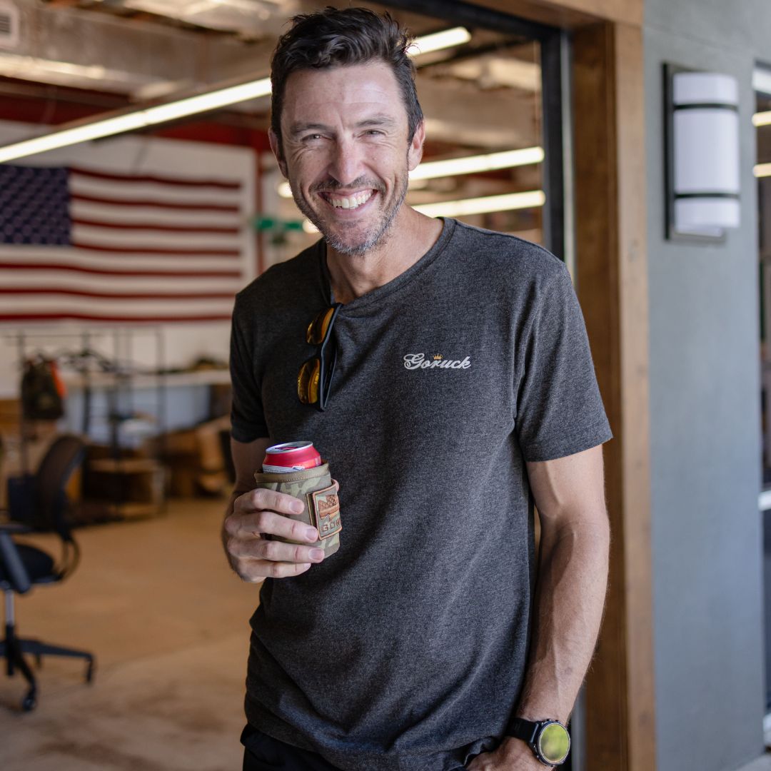 A smiling man wearing a premium Tri-BlendX Goruck t-shirt holds a canned beverage in a camouflage koozie.