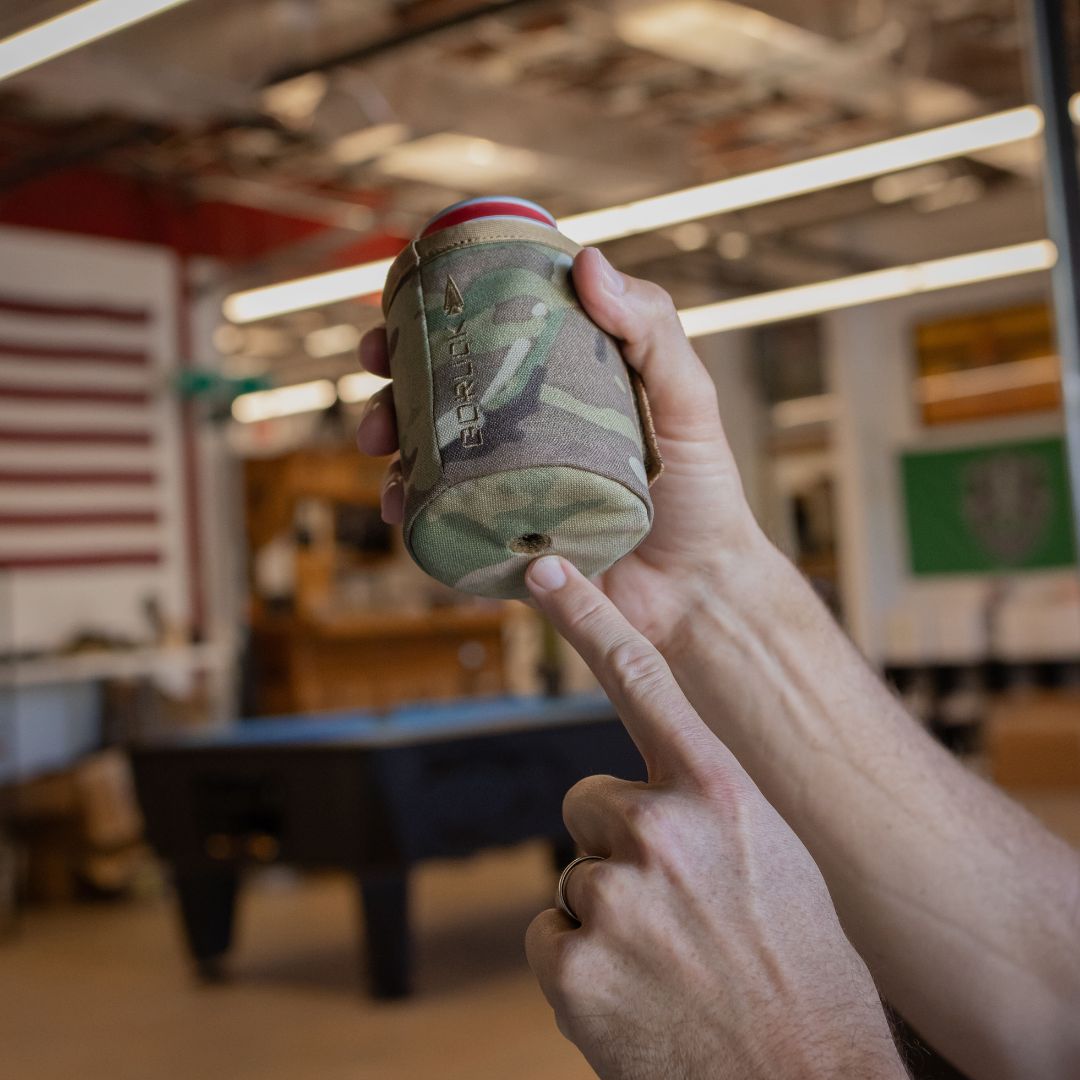 A person points to the drain hole in the bottom of a camouflage-patterned Ballistic Beer Jacket labeled “GORUCK.”