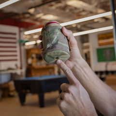 A person points to the drain hole in the bottom of a camouflage-patterned Ballistic Beer Jacket labeled “GORUCK.”